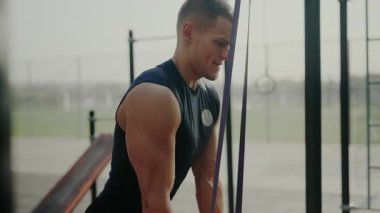 Genç kaslı adam dışarıda direnç bandı eğitimi alıyor. Bu da vücut geliştirme rutininde yoğunluk ve kararlılık gösteriyor. Açık hava Direniş Grubu Eğitim Oturumunda Odaklanmış Sporcu