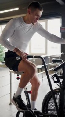 Spor kıyafetleri içindeki formda genç bir adam spor salonunda sabit bir bisikletle çalışıyor, kendini adama ve dayanıklılık gösteriyor. Azimli genç adam yoğun bir şekilde jimnastik bisikleti üzerinde çalışıyor. Kararlı Genç Adam Egzersiz