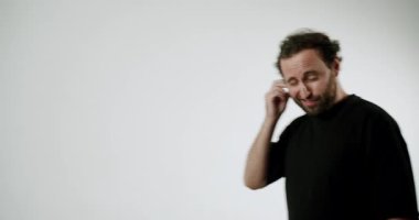 Siyah tişörtlü bir adam kulağını kaşıyarak kameraya göz kırpıyor. Bir jest yapar, dinlediğini söyler ve güler. Basit beyaz bir arkaplanda resmedilmiş..