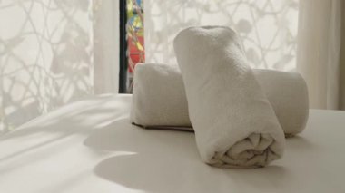 İki kabarık beyaz havlu bir yatağa sarılmış, otel ya da tatil köyündeki misafirler için hazırlar. Güneş ışığı perdelerin arasından akarak huzurlu ve rahatlatıcı bir atmosfer yaratır. seyahat, konukseverlik,