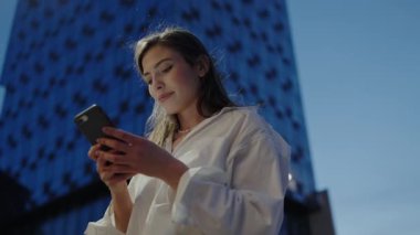 Genç profesyonel bir kadın telefonunu mavi, modern bir binanın önünde dikilirken kontrol ediyor. Teknoloji ve şehir hayatının mükemmel bir karışımı. Akıllı Telefonu Bina Arkaplanına Karşı Kullanan Genç Kadın