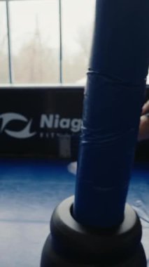 Azimli genç bir adam spor salonunda ağır bir torbaya yumruk atarak yoğun bir şekilde antrenman yapıyor. Gücü, odaklanmayı ve özveriyi yakalar. Spor salonunda boks torbasını yumruklayan odaklanmış erkek sporcu. Dikey video.