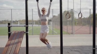 Formda genç bir kadın kollarını kaldırıyor, futbol sahasının yanındaki spor salonuna hazırlanıyor. Görüntü, şehir ortamında bir sağlık ve aktivite hissi yakalıyor..