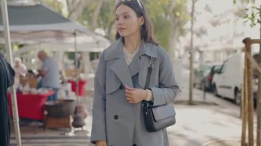 Şık bir trençkot giymiş genç bir kadın, çeşitli tezgahlar ve kentsel yaşamla çevrili, kalabalık bir şehir pazarında düşünceli bir şekilde yürüyor. Genç Kadın Dalgalı Şehir Pazarı 'nda Düşünerek Yürüyor