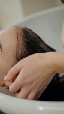 Bir kadın saçını kuaförde yıkatıyor. Kuaför saçına şampuan sürerken nazikçe kafa derisine masaj yapıyor. Bu video rahatlama, şımartma ve kendine bakımı çağrıştırıyor..