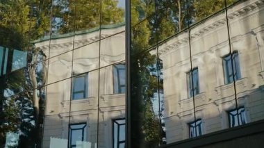 Eski, klasik bir binanın modern bir binanın cam cephesindeki yansıması. Eski ve yeni mimari arasındaki zıtlık gösteriliyor. Güneş ışığı ağaçlardan ve camlardan yansıyor..