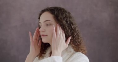 Yüzü sağlıklı cilt masajı elleriyle beyaz bornozlu güzel kadın yüzüne dokunuyor. Yüzüne nazikçe dokunan beyaz bornozlu kadın gözlerinin altından boynuna.