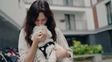 Genç bir kadın küçük beyaz köpeğini nazikçe tutar ve ona sevgi dolu bir öpücük verir. Köpek koşum takımı takıyor ve kollarında rahat görünüyor. Şehir ortamında dışarıdalar.,