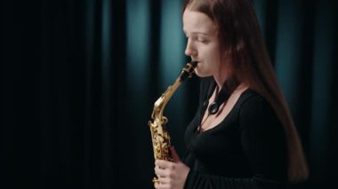 Siyah elbiseli genç bir kadın stüdyoda siyah arka planda saksafon çalıyor. Romantik müzik. Saksafon çalan genç bir bayan müzisyen. Modern stüdyoda saksafon çalan bir kadın..