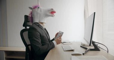 Takım elbiseli, tek boynuzlu at maskeli komik adam mali başarısının tadını çıkarıyor ve işyerinde para sayıyor. Tek boynuzlu at maskeli bir işadamı, yığınla nakit para, dolar kar hesabı yapıyor.