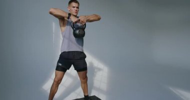 Atletik bir adam, iyi aydınlatılmış bir stüdyo ortamında, dinamik bir idman yapar. Gücünü ve dikkatini gösterir. Stüdyo Ayarlarında Kettlebell ile Genç Atletizm Eğitimi