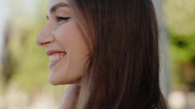 Kahverengi saçlı, gülümseyen genç bir kadının yakın çekimi. Açık havada güneşli bir günün tadını çıkarıyor. Mutluluk ve memnuniyeti ifade ediyor. Gülümseyen kadın doğada güneşli bir günün tadını çıkarıyor.