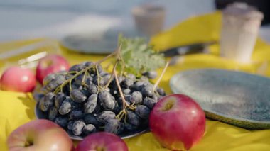 Açık hava pikniğinde canlı sarı bir masa örtüsü üzerinde taze elma ve üzüm tabakları ve deniz manzaralı yakın plan. Taze Elmalar, Üzümler ve Tabaklar Bir Deniz Pikniği İçin Ayarlandı