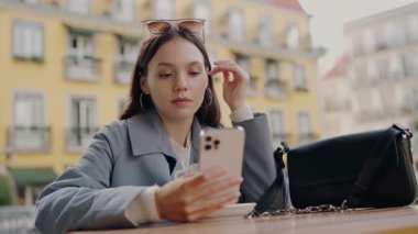 Güneş gözlüğü takan odaklanmış genç bir kadın arka planda renkli binalar bulunan canlı bir şehir bölgesinde otururken akıllı telefonunu kullanıyor. Genç Kadın Renkli Bir Şehirde Telefonunu Kontrol Ediyor