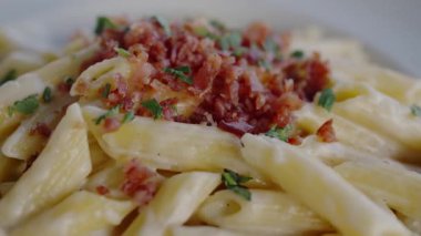 Üzerinde çıtır pastırma parçaları ve taze otlar serpiştirilmiş, beyaz bir kasede servis edilen kremalı makarna yakın çekim görüntüsü. Pastırma parçaları ve taze bitkilerle lezzetli kremalı makarna.