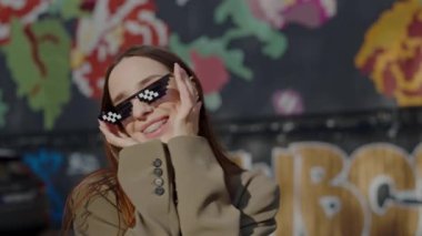 Neşeli genç bir kadın neşeli bir pikselle güneş gözlüğü takıyor ve canlı bir duvar yazısına gülüyor. Şehir tarzı ve gençlik özgürlüğünün mükemmel bir karışımı. Genç Kadın Tarzıyla Açık Hava Eğleniyor