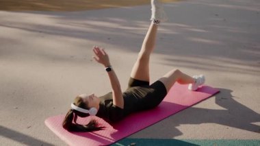 Formda, spor kıyafetli genç bir kadın parkta güneşli bir günün tadını çıkarıyor. Pembe yoga minderinde müzik dinlerken egzersiz yapıyor. Park 'ta Pink Mat' te Açık Hava Sporu Yapan Genç Kadın