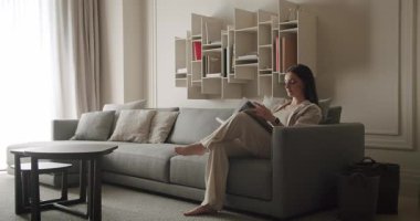 Yumuşak doğal ışık ve minimalist dekorla çevrili, rahat ve modern bir oturma odasında kitap okuyan bir kadın. Modern Salonda Gri Kanepeyle Mobilya Düzenleyen Kadın