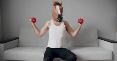 At maskeli komik bir adam evdeki beyaz koltukta ağırlık çalışması için egzersiz yapıyor. Evde komik bir spor salonu var. Komik Sağlık ve Spor Konsepti.