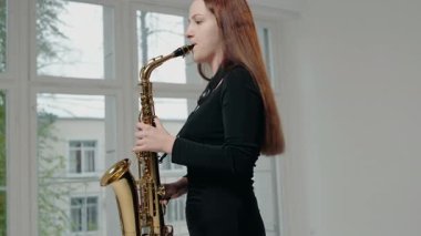 Siyah elbiseli genç bir kadın stüdyoda beyaz arka planda saksafon çalıyor. Saksafon çalan caz müzisyeni. Siyah bir gece elbisesi giymiş, sahnede solo yapan bir kadın saksafoncu..