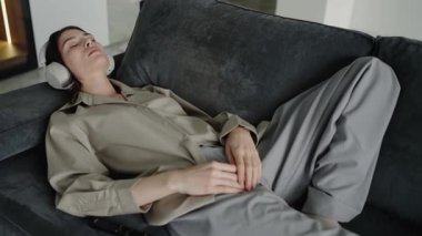 Kanepede uzanmış, kulaklıkla müzik dinleyen genç bir kadın. Sahne sakinlik, sükunet ve rahatlama hissi yayar. Kadın, kulaklıklı koltukta dinleniyor.