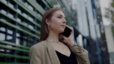 Genç bir bayan profesyonel modern şehir binalarının arka planına karşı cep telefonuyla neşeli bir sohbete girişiyor. Kentsel Ayarlarda Telefonda Konuşan Genç Profesyonel Kadın