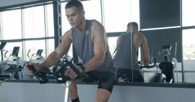İyi donanımlı bir spor salonunda hareketsiz bisiklete binen odaklanmış genç bir adam azim ve fiziksel zindelik sergiliyor. Genç Adam Modern Spor Salonunda Yoğun Bisiklet Çalışmasına Odaklandı