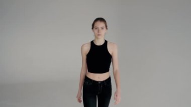 Modaya uygun siyah bir bluz ve kot pantolon giymiş genç bir kadın, modern, minimalist bir stili temsil eden, sade gri bir arkaplan karşısında durmaktadır. Siyah Giyen Genç Kadın