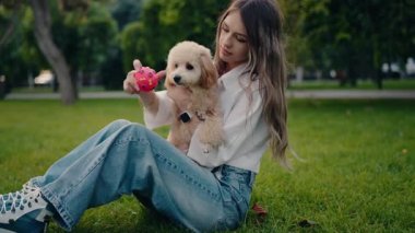 Genç bir kadın parkta çimlerin üzerinde oturuyor, küçük, tüylü bir köpek tutuyor. Ona pembe bir top gösteriyor, yakalamaca oynamaya hazırlanıyor. Köpek sahibinin kollarında mutlu görünüyor. Birlikte güneşli bir günün tadını çıkarıyoruz..