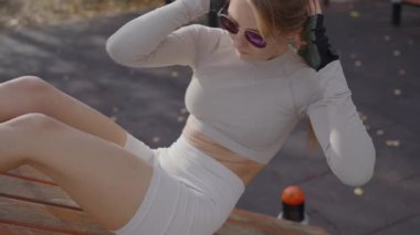 Şık beyaz spor kıyafetleri içinde formda genç bir kadın parktaki bir bankta mekik çekiyor. Açık havada spor ve sağlığın vücut bulmuş hâli. Beyaz Spor 'da Genç Kadın Açık Hava Çalışması sırasında Giyiniyor.