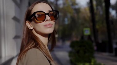 Şık güneş gözlüğü takan genç bir kadın hareketli bir şehir kaldırımında güneşli bir günün tadını çıkarıyor. Moda ve şehir yaşam tarzının mükemmel bir karışımı. Güneş Gözlüklü Zarif Kadın Güneşli Şehir Caddesinde Yürüyor