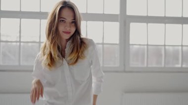 Beyaz gömlek ve kot pantolon giyen genç bir kadın büyük pencereli parlak bir odada güvenle dikiliyor. Onun ifadesi düşünceli ve şık. Beyaz Gömlekli Zarif Genç Kadın