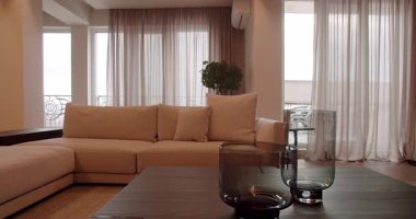 Rahat bej bir kanepe, şeffaf perdeler ve parlak cam bir sehpanın bulunduğu bu modern oturma odasını keşfedin. Konfor ve modernliğin mükemmel bir karışımı. Modern Oturma Odası Doğal Işık