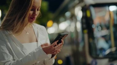 Beyaz elbiseli genç bir kadın, şehir sokaklarında akıllı telefonunu kullanıyor. Şehir ışıklarıyla aydınlatılıyor. Şehirde Gece Akıllı Telefon Kullanan Genç Kadın. Akıllı Telefon Kullanan Genç Kadın.