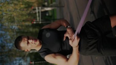Genç, kaslı bir adam, direniş bandıyla açık havada esnemeye yoğunlaşır. Gücünü ve azmini gösterir. Adam dışarıda direniş bandosuyla antrenman yapıyor. Dikey video.