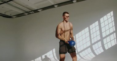23 yaşındaki atletik bir adam parlak ışıklandırılmış bir stüdyoda, fiziksel zindelik ve dayanıklılık eğitimi sergileyen bir su ısıtıcısını kaldırıyor. Güneş Stüdyosunda Kettlebell ile Atletik Genç Adam Eğitimi