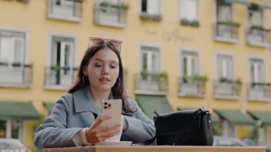 Neşeli genç bir kadın, canlı bir Avrupa şehrinde akıllı telefonunu tutarken, rahat ve mutlu bir şehir yaşam tarzı sergiliyor. Avrupa 'da Akıllı Telefon Kullanırken Genç Kadın El Sallıyor