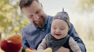 Gülümseyen bir babanın neşeli bebeğini dışarıda renkli bir oyuncakla eğlendirdiği, özel bir ebeveyn-çocuk bağını vurguladığı iç açıcı bir an. Neşeli Baba Park 'ta Gülen Bebekle Oynuyor