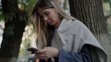 Şehir parkında bir ağacın altında dikilirken akıllı telefonunu karıştıran genç bir bayan. Modern yaşam ve bağlantı konsepti. Urban Park 'ta Ağacın Altında Akıllı Telefon Kullanan Genç Kadın