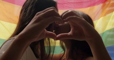 Kadın Lgbt çifti el kalp işareti ile gökkuşağı bayrağının önünde el sallıyor. Lezbiyen aile. Sevgi ve desteği paylaşan iki kadın el ele tutuşuyor. Yaşam tarzı ve ilişkiler. LGBT kavramı