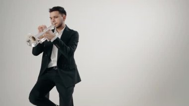 Takım elbiseli genç ve yakışıklı bir adam arzu ve ilhamla trompet çalar. Modern bir stüdyoda beyaz arka planda trompet çalan bir erkek caz şarkıcısı..