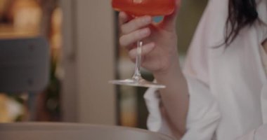 Zarif bir şekilde tasarlanmış bir bardakta canlı turuncu bir kokteyli zarif bir şekilde tutan şık bir kadının yakın çekim görüntüsü. Bulanık arka plan samimi ve samimi bir atmosferi işaret ediyor.