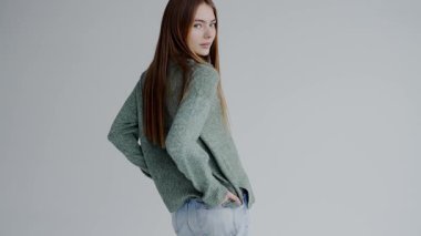 Yeşil kazak ve kot pantolon giyen uzun saçlı genç bir kadın gri arka planda rahat ve kendinden emin bir görünüm sergiliyor. Yeşil Kazaklı Genç Kadın Stüdyoda Gelişigüzel poz veriyor