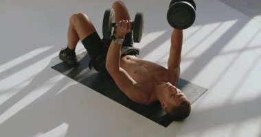 23 yaşında atletik yapılı bir adam, iyi ışıklandırılmış bir stüdyoda, minder üzerinde ağırlık çalışması yapıyor. Yoğun Spor Salonu Eğitimine Başlayan Genç Atletik Adam