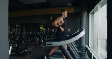 Formda bir genç adam, iyi donanımlı bir spor salonundaki koşu bandında koşuyor, odak ve kararlılığını ifade ediyor, doğal ışığın mekanı aydınlattığı çağdaş bir ortamda..