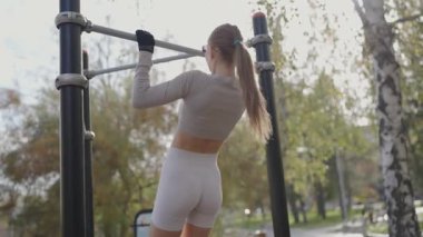 Atletik giysiler içindeki odaklanmış bir kadın güneşli bir parkta barfiks çekerek güç ve zindelik gösterisi yapıyor. Fitness Coşkusu Park 'taki Spor Salonu' nda barfiks çekiyor