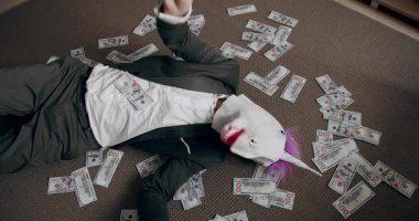 Tek boynuzlu at maskeli komik bir adam mali başarısının tadını çıkarıyor ve yerde oturmuş paraları sayıyor. Tek boynuzlu at maskeli adam para atıyor. Para yağmuru, düşen dolarlar. Yavaş çekim.