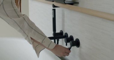 Çağdaş bir banyodaki küvetin siyah ve modern musluğunu açıyor. Su beyaz küvete akar. Bu şık banyo eşsiz bir fayans tasarımı ve ahşap bir raf içeriyor..