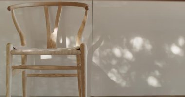 Doğal ışık yaprakların arasından süzülür, örülmüş ahşap koltuklu şık bir sandalyenin arkasındaki minimalist beyaz mutfak dolabının üzerine dans eden gölgeler bırakır. Sahne bir huzur ve sükunet hissi uyandırıyor.,