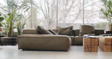Modern oturma odasının içinde büyük bir deri kanepe, ahşap sehpa ve pencere kenarında yemyeşil bitkiler var. Doğal ışık uzayı aydınlatır, rahat ve çağdaş bir atmosfer yaratır..
